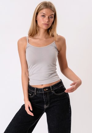Jeune femme portant un débardeur gris clair et un jean noir, debout avec une main près de sa taille sur un fond blanc uni.