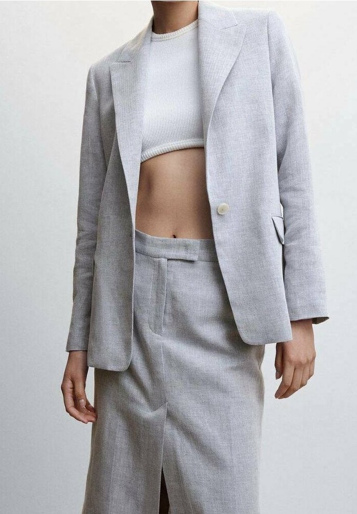 Femme portant un blazer ajusté gris clair et une jupe taille haute assortie avec une fente à l'avant, associée à un top court blanc côtelé.