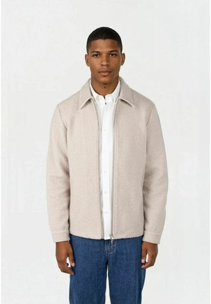 Mann iført beige fleecejakke med glidelås over hvit skjorte med knapper og blå jeans, stående mot en ensfarget lys bakgrunn.