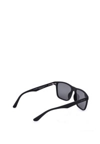 Lunettes de soleil rectangulaires noires avec verres foncés et branches fines texturées reposant ouvertes sur une surface blanche.