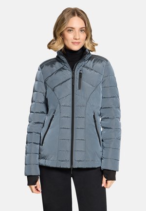 Woman wearing a blue quilted jacket with zipped pockets and black inner sleeves, standing against a plain light background.