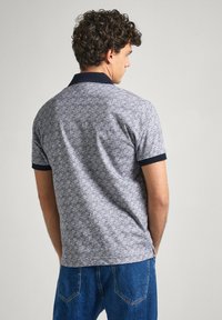 Polo tričko s námořnickým límcem a modrobílým geometrickým vzorem, krátké rukávy a rovný lem. Tkanina vypadá texturovaně a prodyšně.