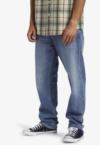 Jeans de mezclilla azul claro con un corte recto, usados con una camisa de cuadros en verde y beige y unas zapatillas de lona negras.