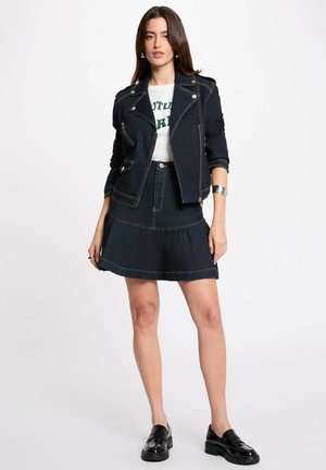 Donna con giacca di jeans scura e gonna abbinata, T-shirt bianca stampata, mocassini neri e bracciale a polsino argento, in piedi davanti a uno sfondo bianco.