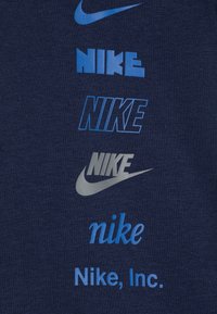 Tamsiai mėlynas audinys turi kelis „Nike“ logotipus skirtingose mėlynos ir sidabrinės atspalviuose, pateikdamas įvairius šriftų stilius ir dydžius.