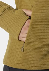 Chaqueta de forro polar texturizado de color mostaza, con un bolsillo con cremallera, puños ajustados y un corte ceñido en la cintura, que enfatiza la comodidad y el calor.