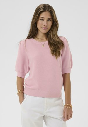 Młoda kobieta o długich brązowych włosach, ubrana w różowy sweter z krótkim rękawem, białe spodnie i złotą biżuterię, stojąca z rękami w kieszeniach.