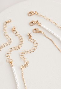 Goldtone jewelry chains with varying links and textures, including pearl accents and lobster clasps, arranged on a light background.