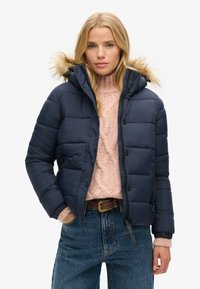 Veste matelassée marine avec capuche en fourrure, design matelassé, fermeture éclair et boutons-pression, portée sur un pull en tricot rose pâle.