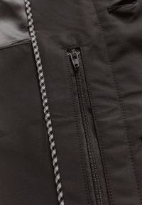 Czarna kurtka na zamek z gładką fakturą; posiada wzorzysty suwak i kontrastowe przeszycia wzdłuż linii zamka.