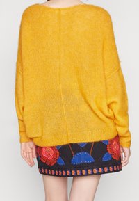 Överstor, gul, ullblandad tröja med en mjuk textur, tillsammans med en mönstrad svart kjol som har blå och röda blommönster broderade.