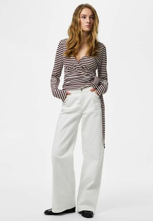 Jeune femme aux longs cheveux ondulés, portant un haut portefeuille rayé marron et blanc et un pantalon blanc à jambes larges, debout les mains dans les poches.