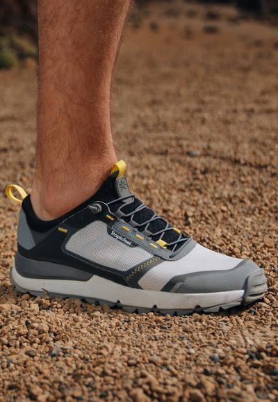 Sport shoe with a grey, white, and black mesh upper, yellow accents, and a rugged rubber sole. Features a reinforced toe and elastic laces.