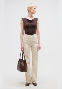 Γυναίκα με κοντά ξανθά μαλλιά, που φοράει ένα αμάνικο καφέ τοπ, μπεζ ψηλόμεσα παντελόνια, καφέ παπούτσια και κρατά μια μεγάλη καφέ τσάντα.