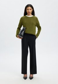 Olivno zelena pletena majica z belim ovratnikom, kombinirana z visokim pasom črnih hlač in črnimi koničastimi petami. V roki drži torbico za v roko.