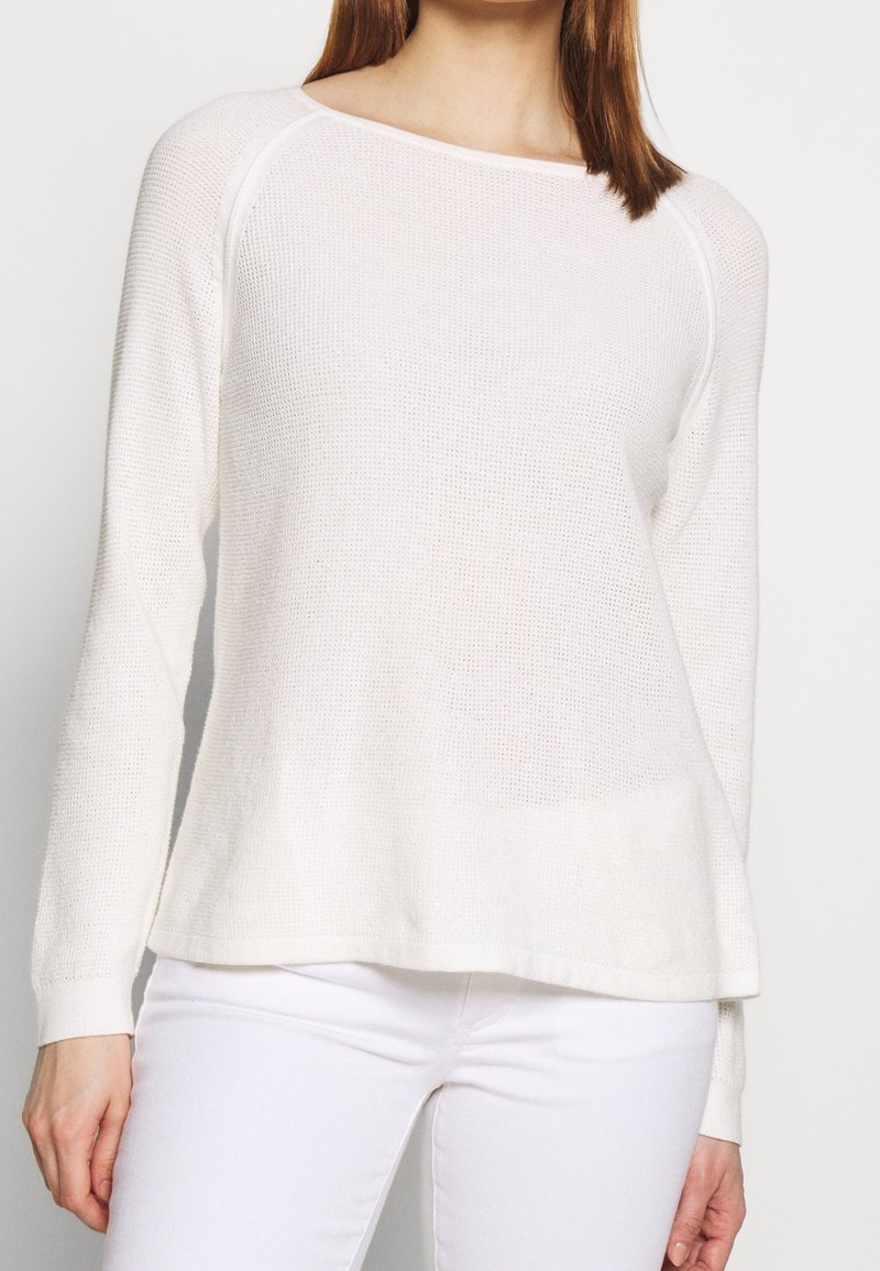 Femme portant un pull en maille blanc à manches longues et un pantalon blanc, debout devant un fond clair uni.