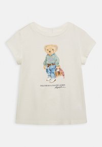 Camiseta de manga corta color crema con una ilustración de un oso que lleva un suéter verde, jeans azules y sostiene una pequeña bolsa con acentos rosas.