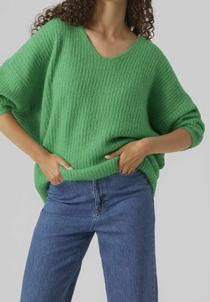 Femme portant un pull vert texturé ample à manches trois-quarts et un jean denim bleu, les mains glissées dans les poches avant.
