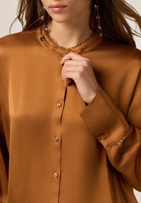 Brązowa satynowa koszula o miękkiej fakturze, z zapięciem na guziki z przodu i szydełkowym obszyciem przy kołnierzu. Długie rękawy z guzikami jako akcent.