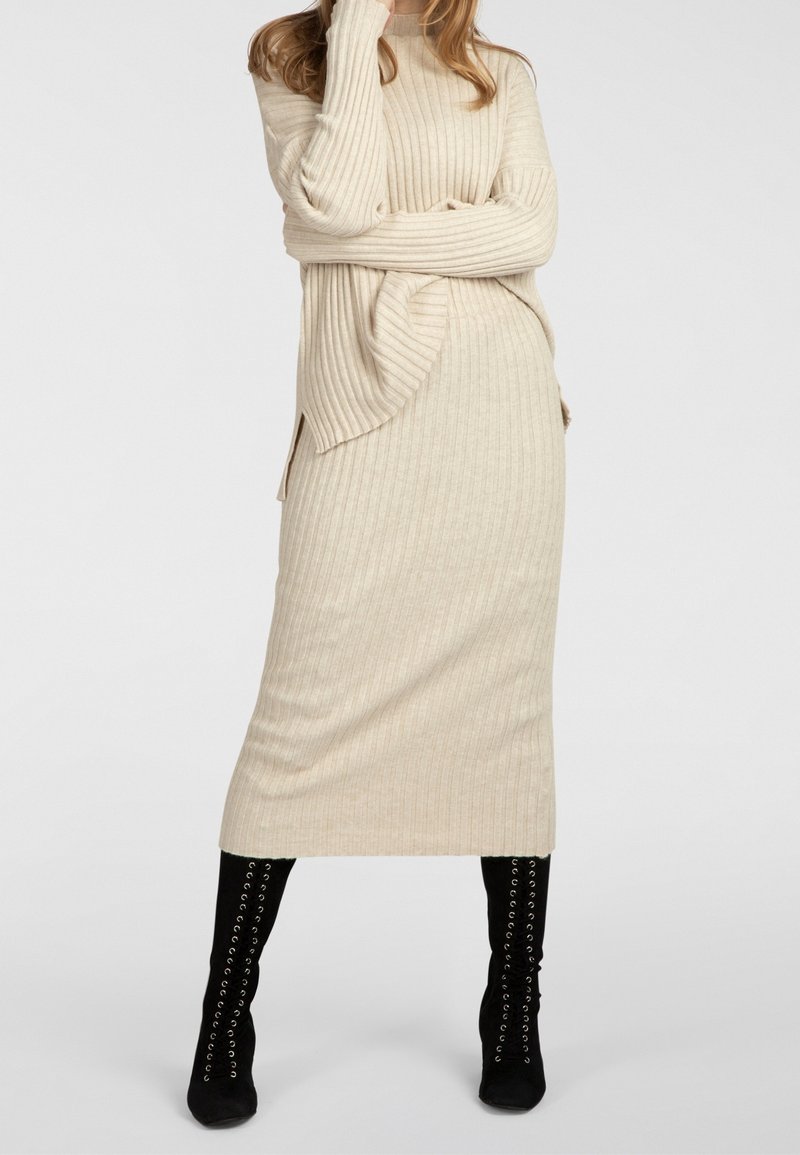 Femme portant un pull en tricot côtelé beige et une jupe midi assortie, associée à des bottes noires à lacets montant jusqu'aux genoux, debout les bras croisés.