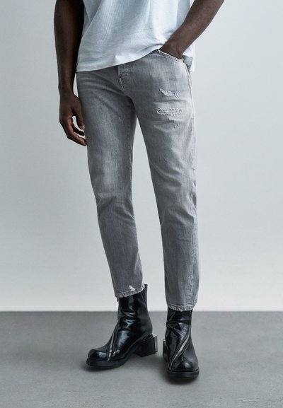 Homme portant un jean gris clair usé associé à des bottines en cuir noir à fermeture éclair frontale, debout sur un sol gris devant un fond uni.
