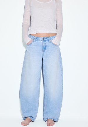 Osoba stojaca bosá na bielom pozadí, oblečená v voľných svetlomodrých džínsach s širokými nohavicami a priehľadnom bielej blúzke s dlhými rukávmi.