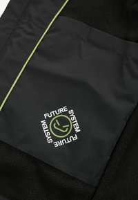 Veste noire en texture polaire, dotée d'un contour vert et d'un logo circulaire avec le texte "FUTURE SYSTEM" en blanc.
