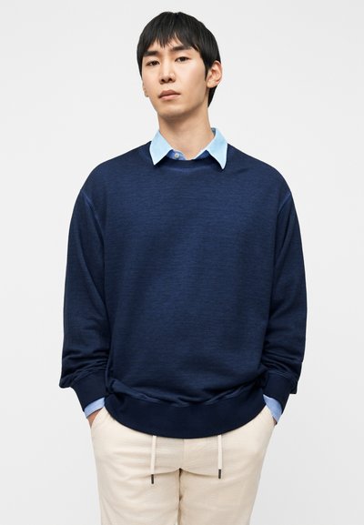 Φούτερ σε ναυτικό μπλε με υφή, μακριά μανίκια και στρογγυλή λαιμόκοψη, συνδυασμένο με ανοιχτό μπλε πουκάμισο με γιακά από κάτω. Μπεζ παντελόνι με κορδόνι στη μέση.