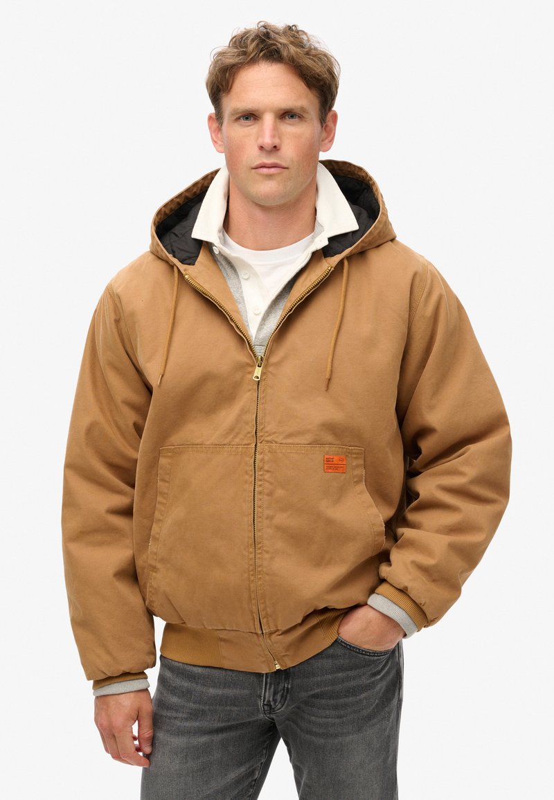 Chaqueta con capucha marrón y cremallera, con un gran bolsillo frontal, puños acanalados y un forro interno en contraste. Llevada sobre una camisa blanca.