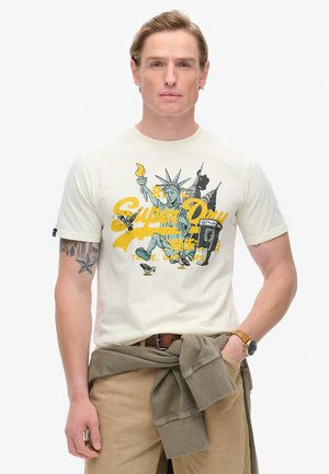 Camiseta crema con un gráfico colorido de la Estatua de la Libertad, pájaros y texto, combinada con pantalones beige y un suéter verde atado a la cintura.
