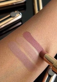 Matte leppestift i tre swatcher på en hudtonet overflate, som spenner fra lys mauve til dypere plomme, med en leppestift i gull-akenter.