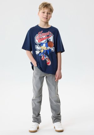 Boy with short blond hair stands wearing blue Sonic the Hedgehog basketball graphic t-shirt, gray jeans, and white sneakers on a white background.