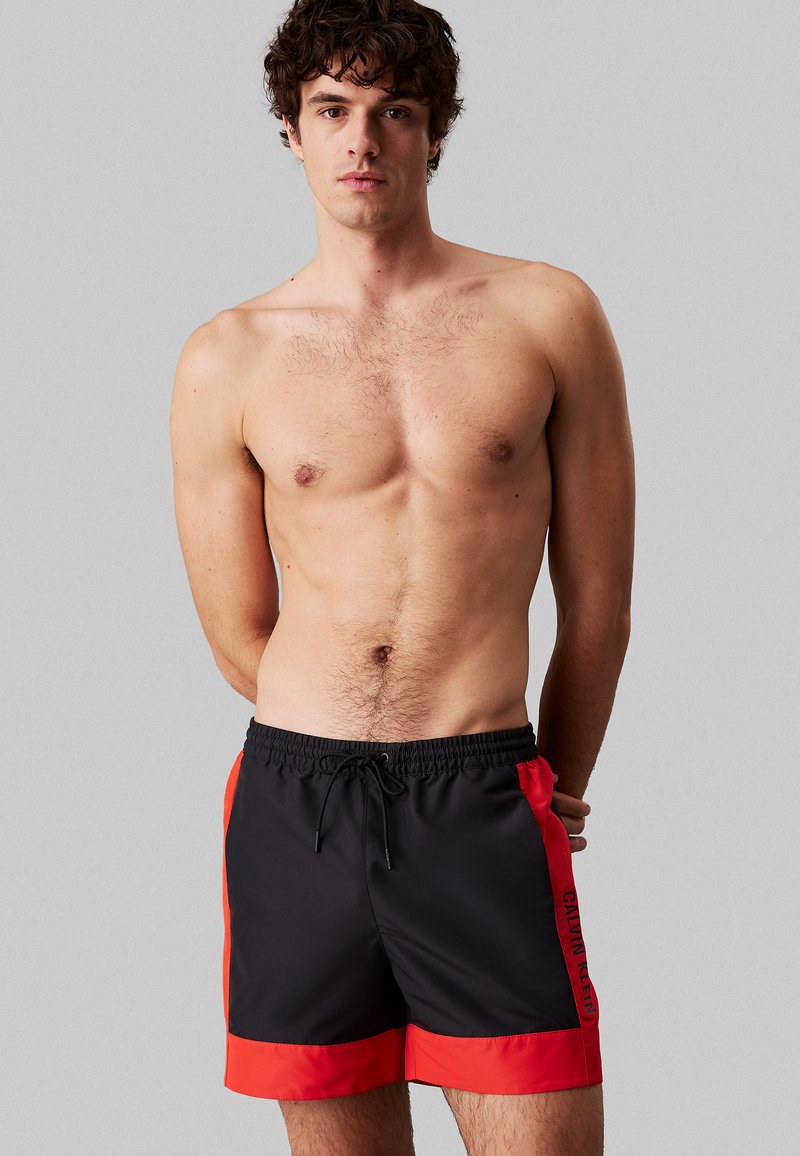 Costume da bagno maschile nero con pannelli laterali e dettagli rossi. Vita elastica con cordino. Realizzati in materiale leggero e ad asciugatura rapida.