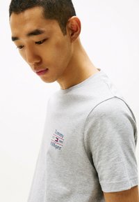 Camiseta de algodón gris con cuello redondo y mangas cortas; logo bordado "Tommy Hilfiger" con acentos rojos y azules en el pecho.