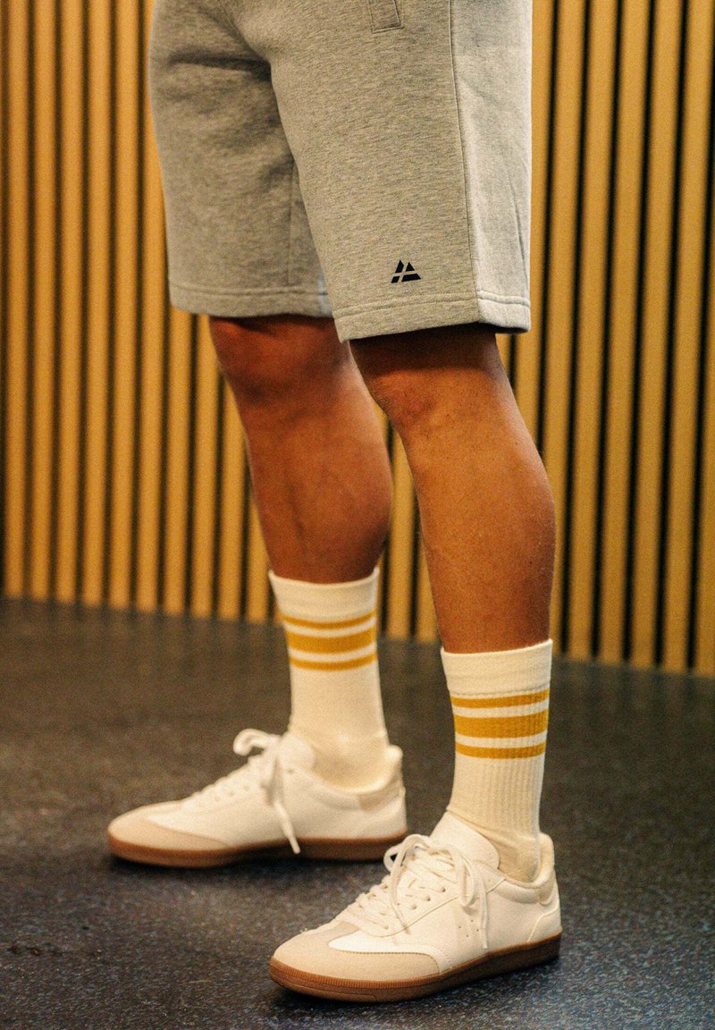 Grå sportshorts med en svart logotyp, matchat med vita strumpor med två gula ränder och vita sneakers med en brun sula.