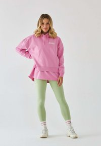 Przesadny różowy hoodie z kieszenią z przodu i dopasowaną różową warstwą podstawową, zestawiony z jasnoróżowymi legginsami i białymi sneakersami z różowymi akcentami.