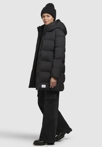 Jachetă puffer neagră cu glugă, dotată cu închidere prin fermoar și nasturi, design matlasat și emblemă pe partea inferioară frontală; purtată peste pantaloni negri largi.