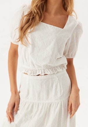 Biała bluzka z haftowanego materiału z krótkimi rękawami i falbanką w pasie, w połączeniu z dopasowaną plisowaną spódnicą, noszona przez osobę z rozpuszczonymi włosami.