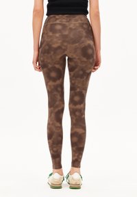 Vrouw gezien vanaf de achterkant, gekleed in strakke bruine legging met een patroon, in combinatie met casual witte en groene sneakers, tegen een effen achtergrond.