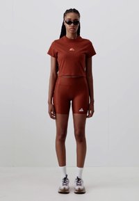 Rust-colored short-sleeve top and bike shorts with logo. Fitted design, smooth texture. Model wears black sunglasses and sneakers with white socks.