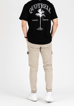 Mężczyzna w czarnej koszulce z białą palmą i napisem, beżowych spodniach typu cargo oraz białych trampkach, stoi tyłem z rękami w kieszeniach.