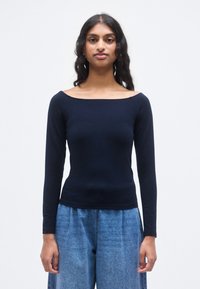 Μπλε σκούρο μπλουζάκι μακρυμάνικο με ντεκολτέ που αφήνει τους ώμους ακάλυπτους, εφαρμοστή γραμμή και απαλή υφή, συνδυασμένο με ψηλόμεσο ανοιχτό μπλε τζιν παντελόνι.