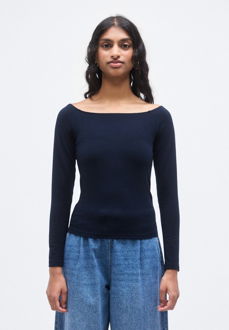 Μπλε σκούρο μπλουζάκι μακρυμάνικο με ντεκολτέ που αφήνει τους ώμους ακάλυπτους, εφαρμοστή γραμμή και απαλή υφή, συνδυασμένο με ψηλόμεσο ανοιχτό μπλε τζιν παντελόνι.