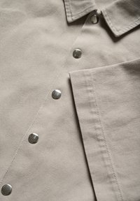 Ljust beige, texturerad tyg med en knappslå med silverdetaljer och en vikt del som avslöjar den inre strukturen.