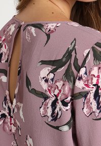 Blommig blus i mjuk mauve-nyans med detalj i form av en nyckelhål i ryggen, prydd med rosa och mörkgröna blommönster samt knappstängning.