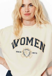 Femme blonde portant un sweatshirt crème avec le texte "FEMMES" et "Depuis 1975", posant les mains sur les hanches devant un fond blanc.