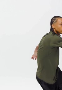 Dlhé rukávy, olivovo zelená košeľa s voľným strihom, so zaobleným lemom a bočnými rozparkami; skombinovaná s čiernymi nohavicami.