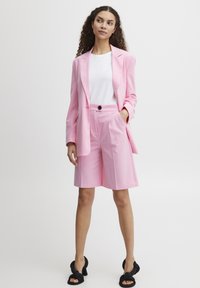 Roze suitjacket en bijpassende shorts, gladde stof, op maat gemaakt ontwerp, mouwlengte tot de elleboog, zwarte knoopdetails, gecombineerd met een wit topje.