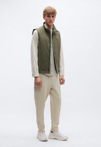 Bezrękawnik w oliwkowej zieleni na białej koszuli w paski, zestawiony z jasnobeżowymi spodniami i jasnymi sneakersami, pokazany na jednolitym tle.