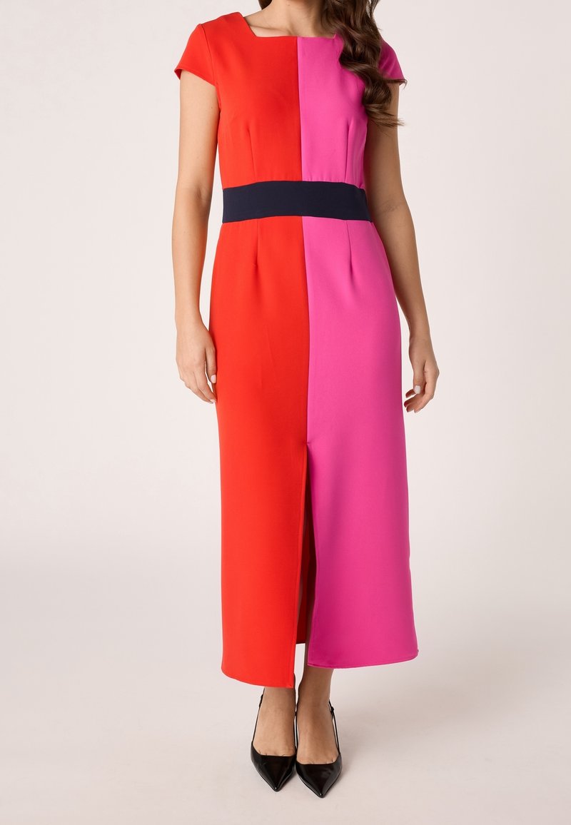 Femme portant une robe color block avec côté gauche rouge, côté droit rose, ceinture noire, manches courtes, fente sur le devant et talons noirs pointus.